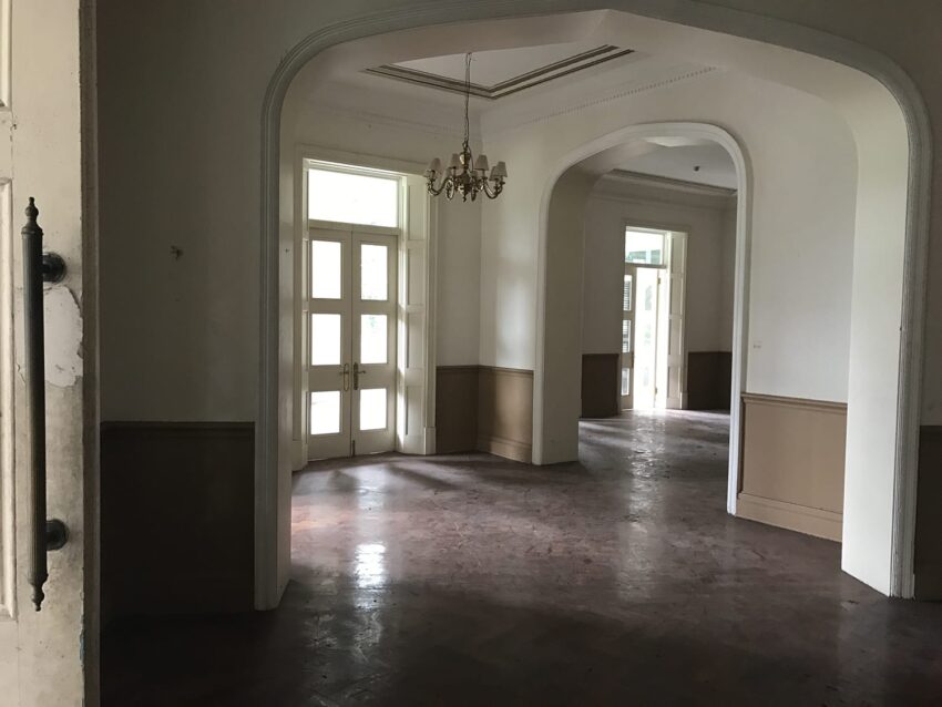 Spacious, empty room with large windows, chandelier, and archways, featuring a wooden floor and beige and white walls.