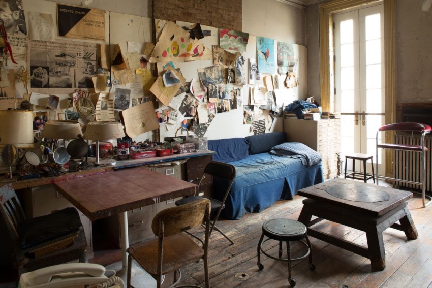 Cozy artist's studio with a couch, wooden tables, chairs, and walls covered in posters and sketches near a sunlit window.