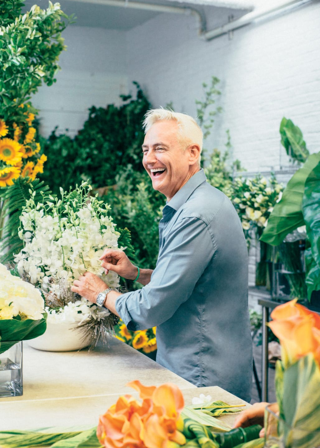 A man smiling and arranging flowers in a bright floral shop with various colorful blooms around him.