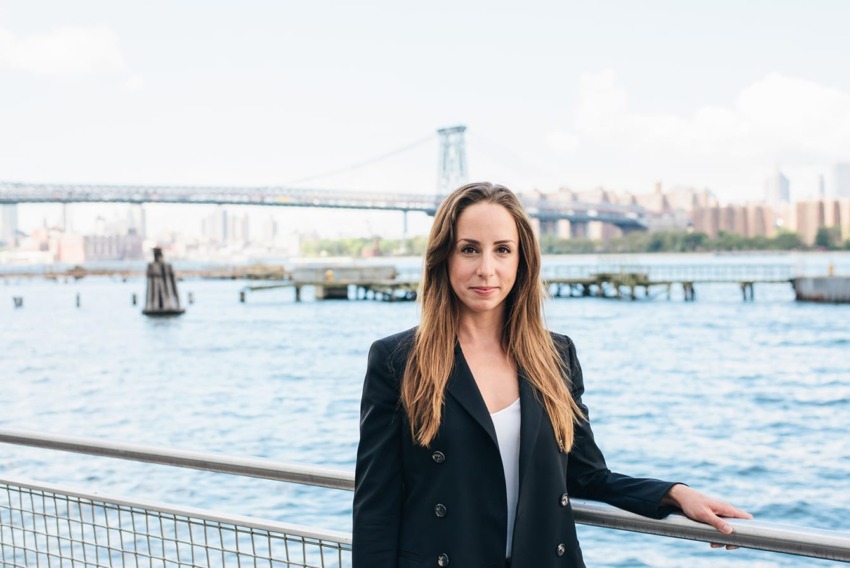 Person standing by a railing with a river and bridge in the background on a sunny day.