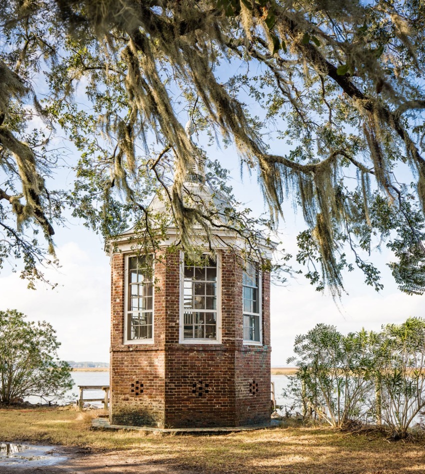 Historic brick gazebo under Spanish moss-draped trees by a lake on a sunny day.