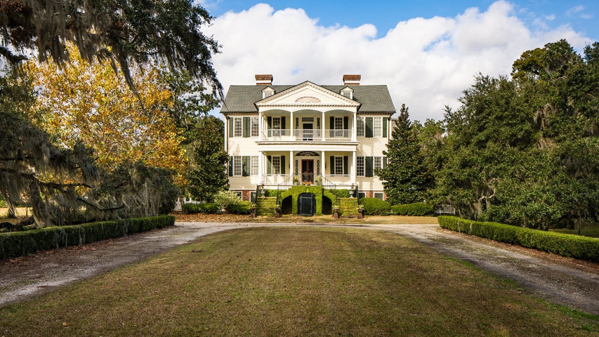 Historic colonial-style mansion surrounded by trees and a spacious lawn under a partly cloudy sky.
