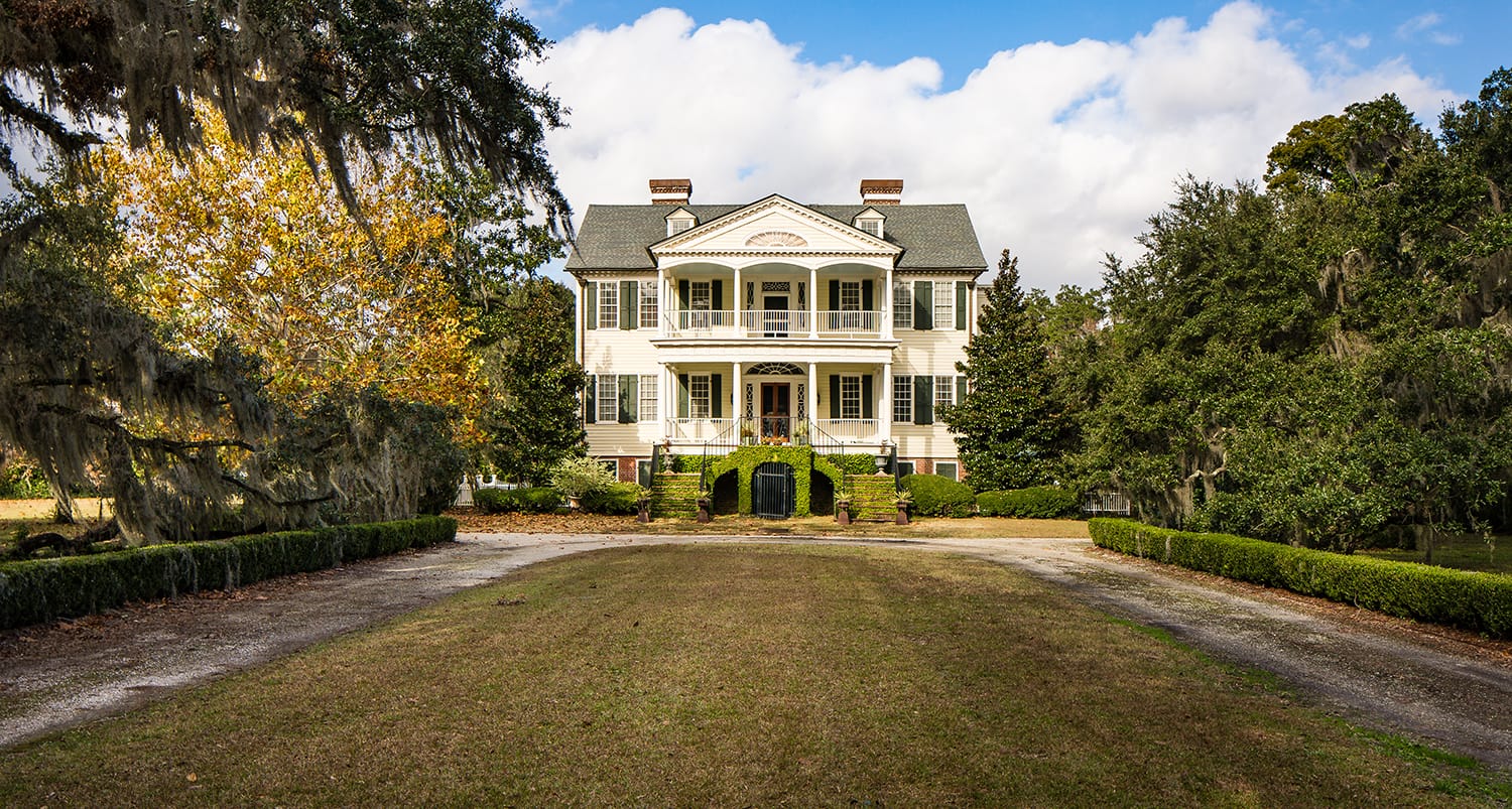 Historic colonial-style mansion surrounded by trees and a spacious lawn under a partly cloudy sky.