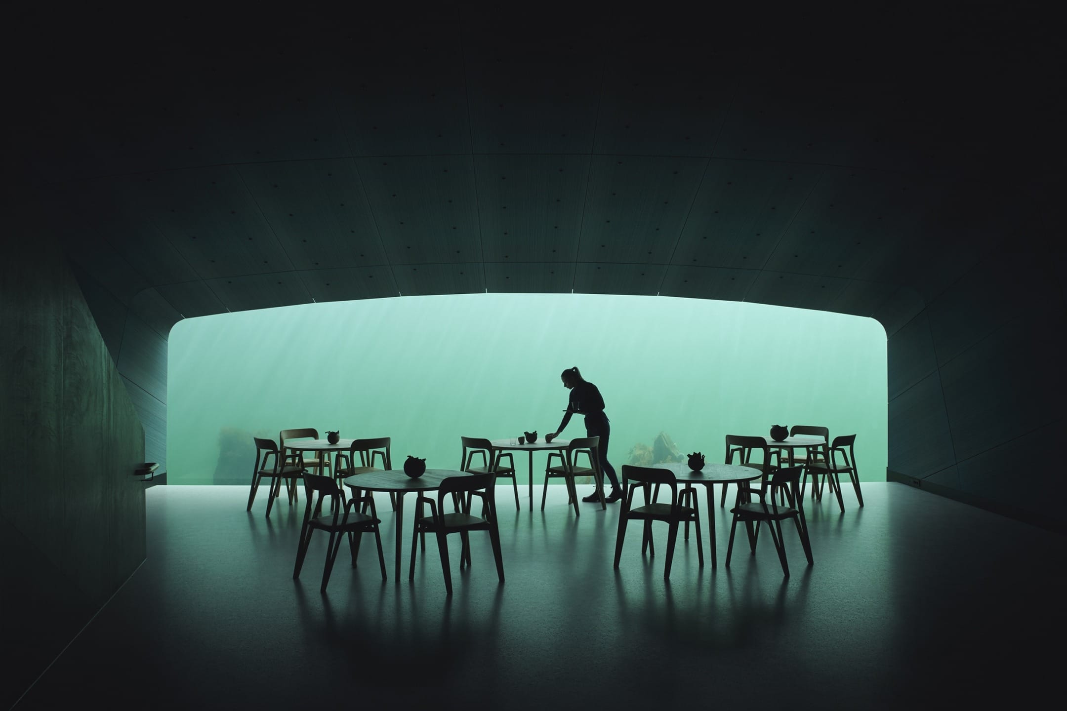 Silhouette of a person arranging tables in a dimly lit underwater restaurant with large window view of ocean.