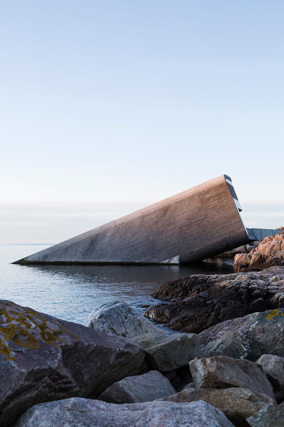 Angled concrete structure at the water's edge against a clear sky, surrounded by mossy rocks and calm sea.