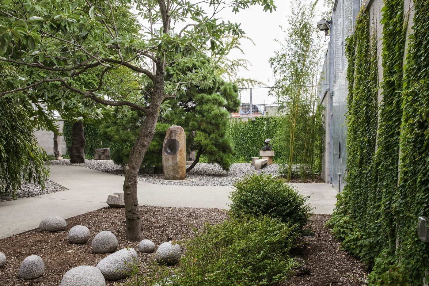 Zen garden with stone sculptures, lush trees, and gravel pathways surrounded by green ivy-covered walls.