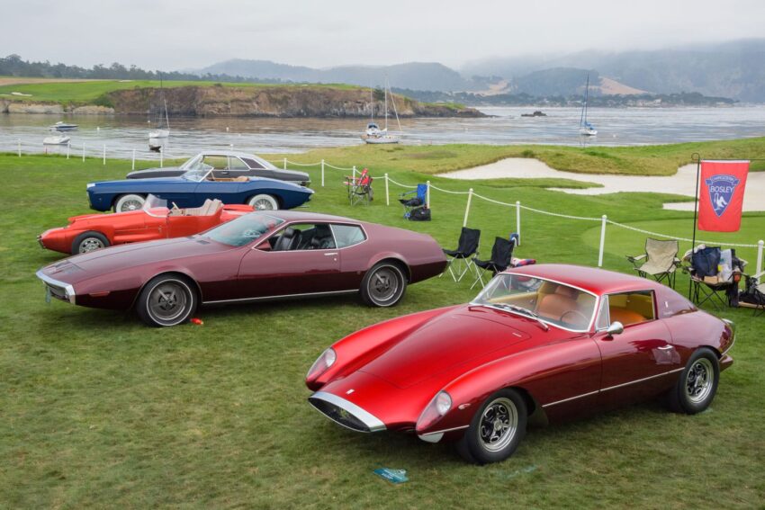 Classic cars displayed on a grassy field by the ocean, with chairs and a banner nearby.