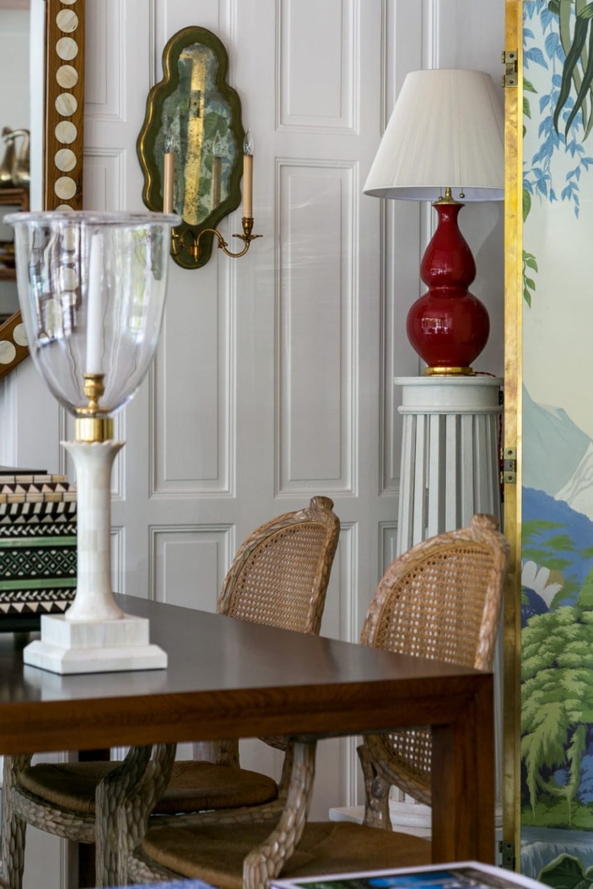 Elegant dining room with wicker chairs, wooden table, red lamp on stand, wall sconce, and decorative mirror.