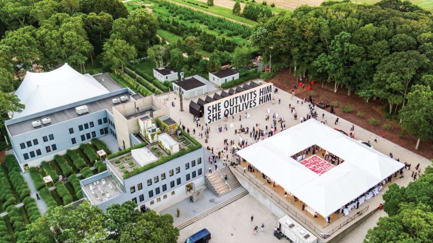 Aerial view of a modern art complex surrounded by trees, with people gathered near a large outdoor tent and bold text art display.