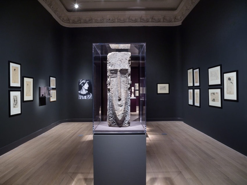 Art gallery interior with large stone sculpture on display, surrounded by framed sketches on dark walls.