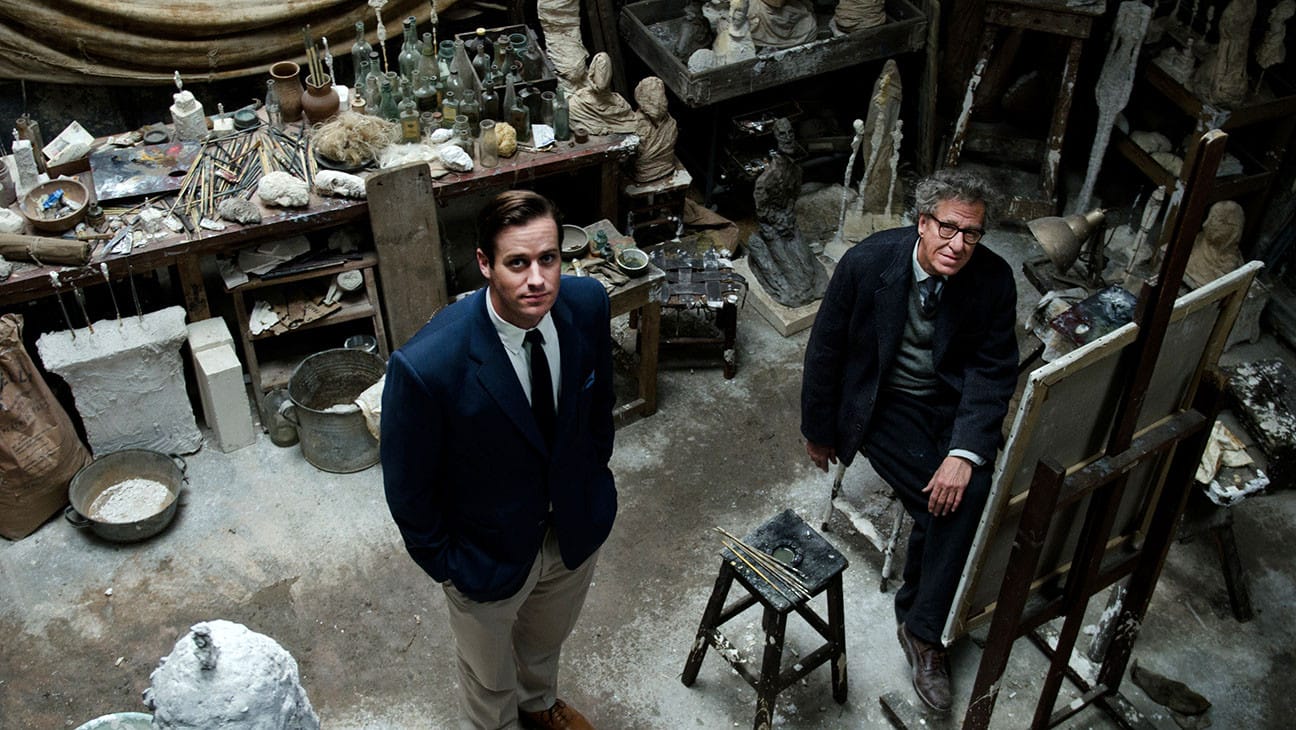 Two men in a cluttered artist studio surrounded by sculptures, easels, and art supplies, one seated and one standing.