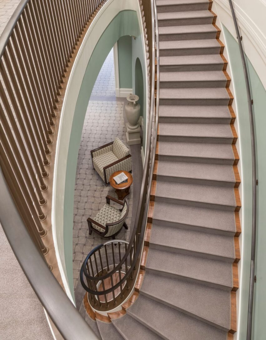Curved staircase with carpeted steps, ornate railing, and chairs with a table in an elegant, light-filled interior space.