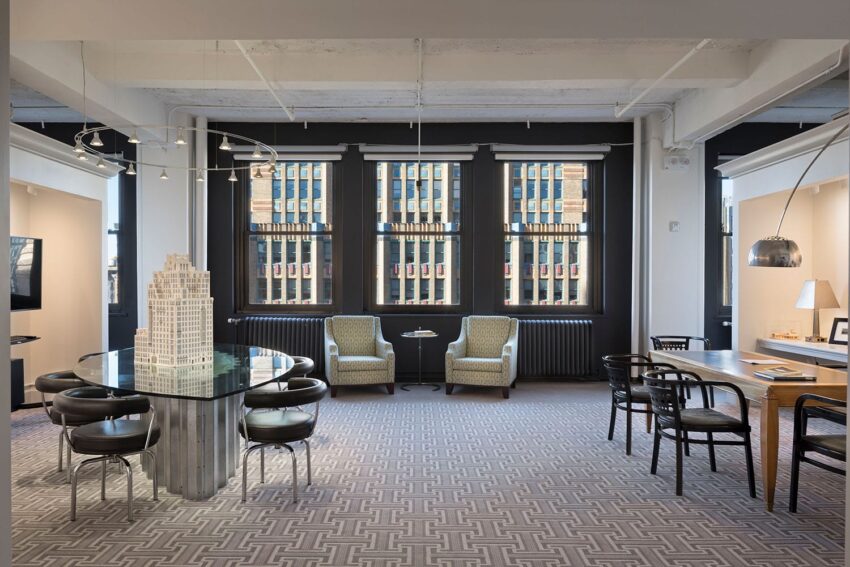 Modern office space with a round table, chairs, and large windows overlooking a cityscape, featuring neutral-toned decor.