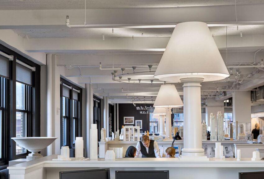 Modern architectural studio interior with large lamp fixtures, models on display, and two people working at desks.