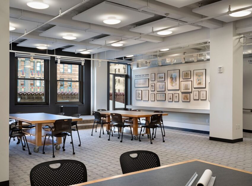 Modern study room with tables, chairs, large windows, framed artwork on walls, and ceiling lights.