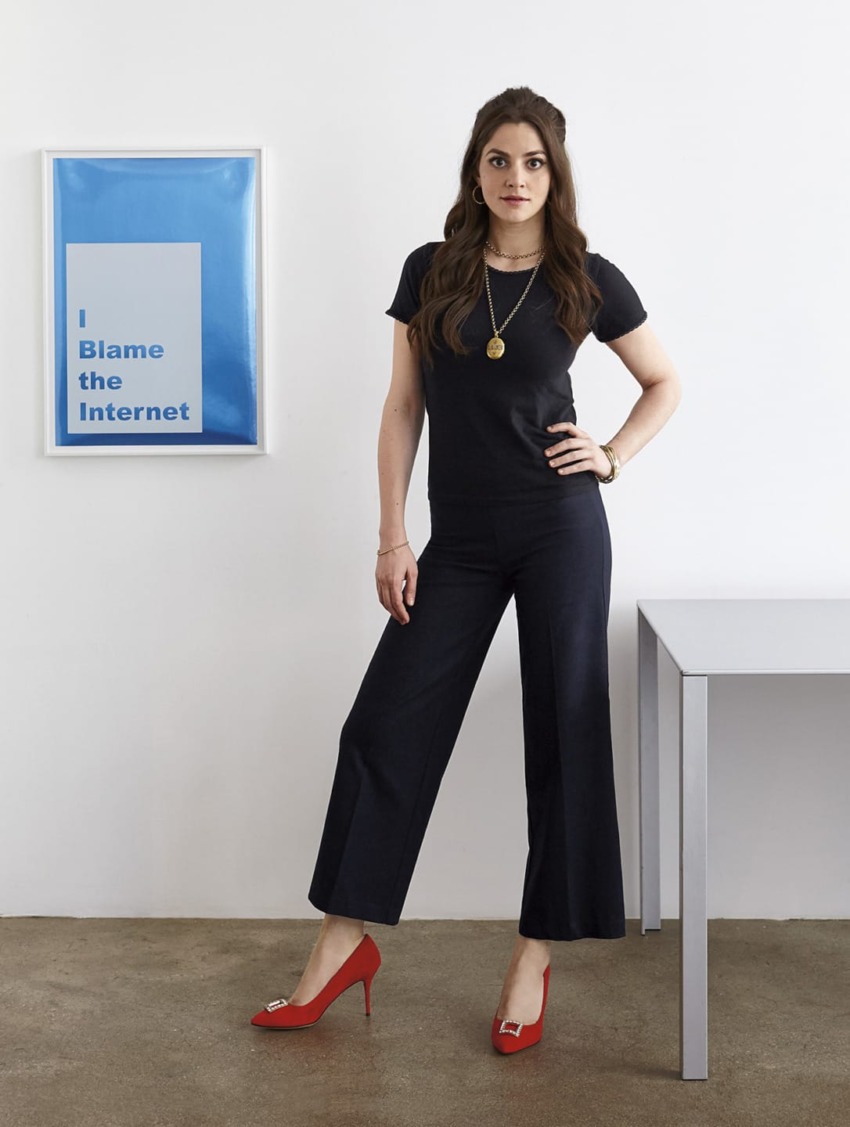 Woman in black outfit, red heels stands next to a desk with a framed blue poster stating "I Blame the Internet" on the wall.