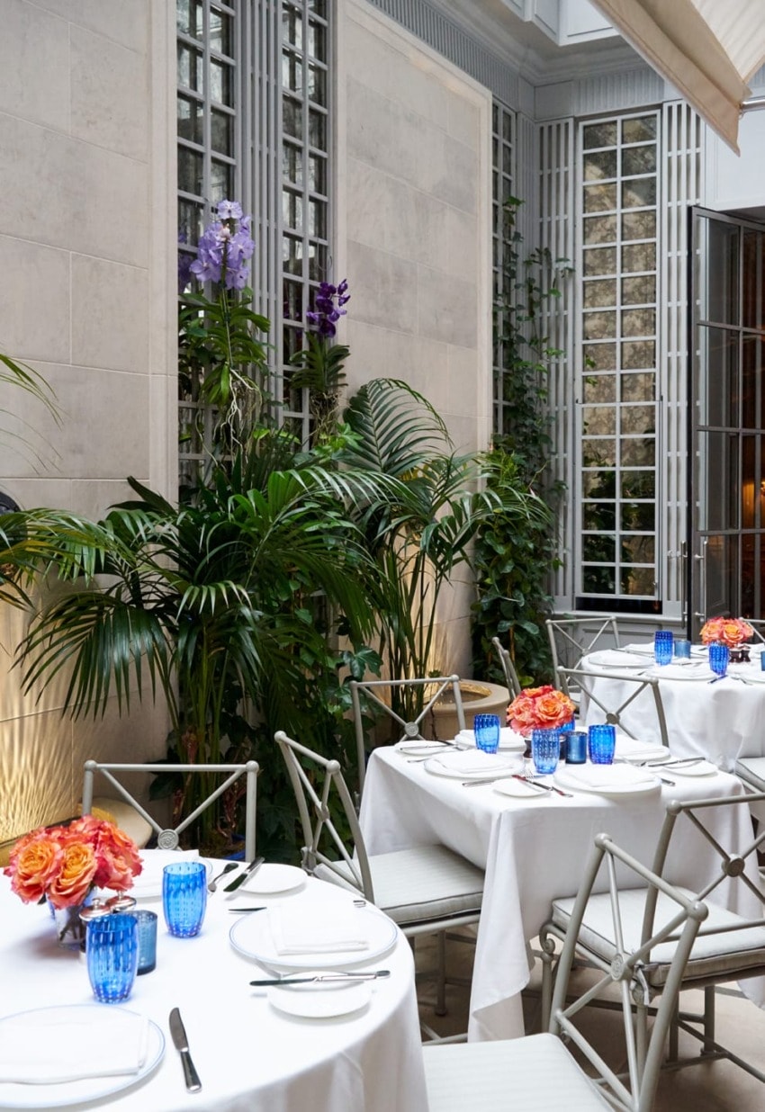 Elegant restaurant dining area with white tablecloths, vibrant flowers, blue glassware, and lush green plants along the walls.