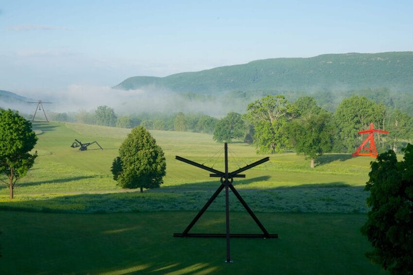 Landscape with large metal sculptures in a grassy field, surrounded by trees and mountains under a clear blue sky.