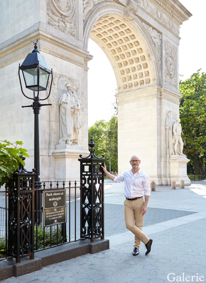 Person standing near a black gate and a historic arch in a sunlit park setting.
