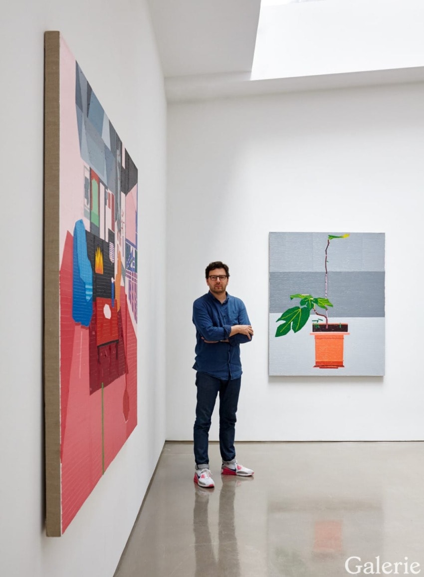 Man standing in art gallery with colorful abstract paintings on walls, wearing blue shirt and jeans.
