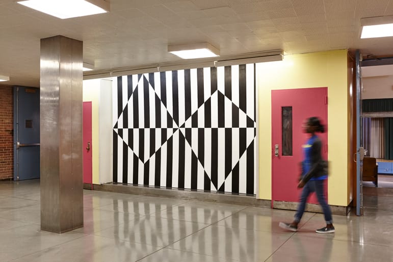Person walking past a black and white geometric wall mural with pink doors on either side in a modern hallway.