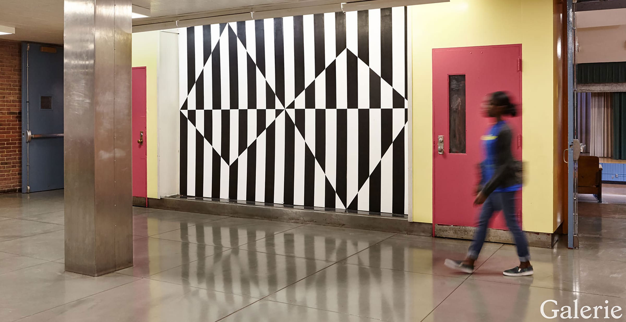 Person walking in gallery with geometric black and white artwork on the wall, pink doors adjacent.