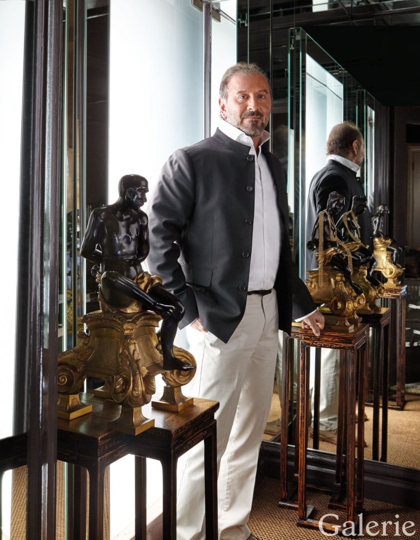 Man in formal attire standing next to decorative sculptures in a stylish room with mirrors and elegant decor accessories.
