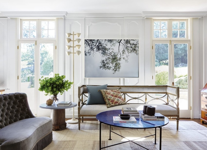 Modern living room with large windows, stylish furniture, abstract wall art, and lush greenery in a cozy, bright setting.
