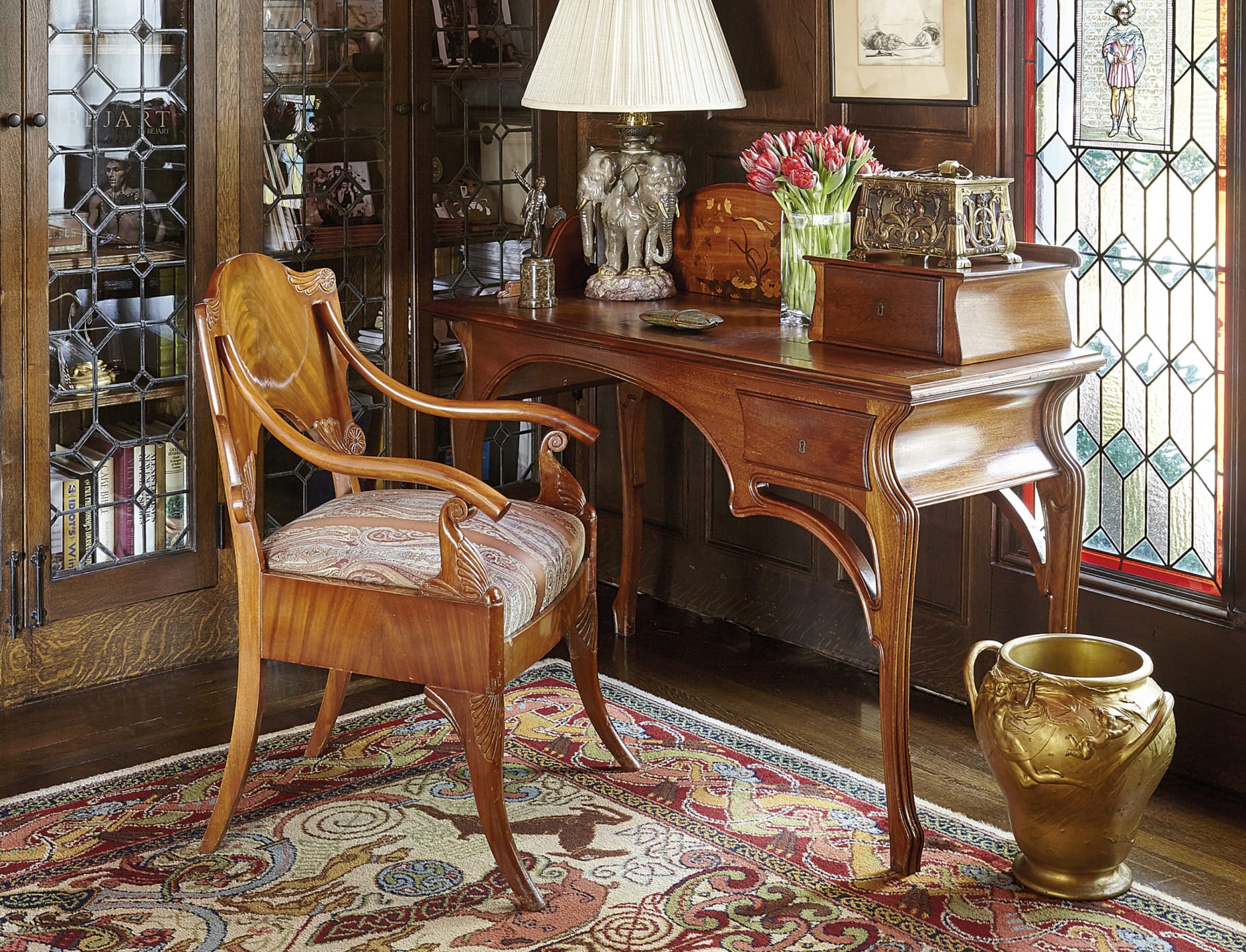 Antique wooden desk and chair in a cozy room with ornate decor, stained glass window, lamp, and vases on patterned rug.