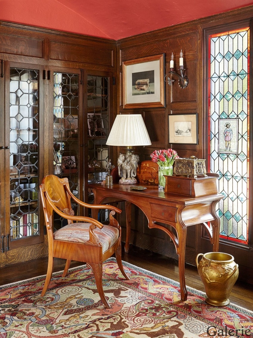 Vintage study with wooden desk, ornate chair, stained glass window, floral carpet, table lamp, and decorative vases.