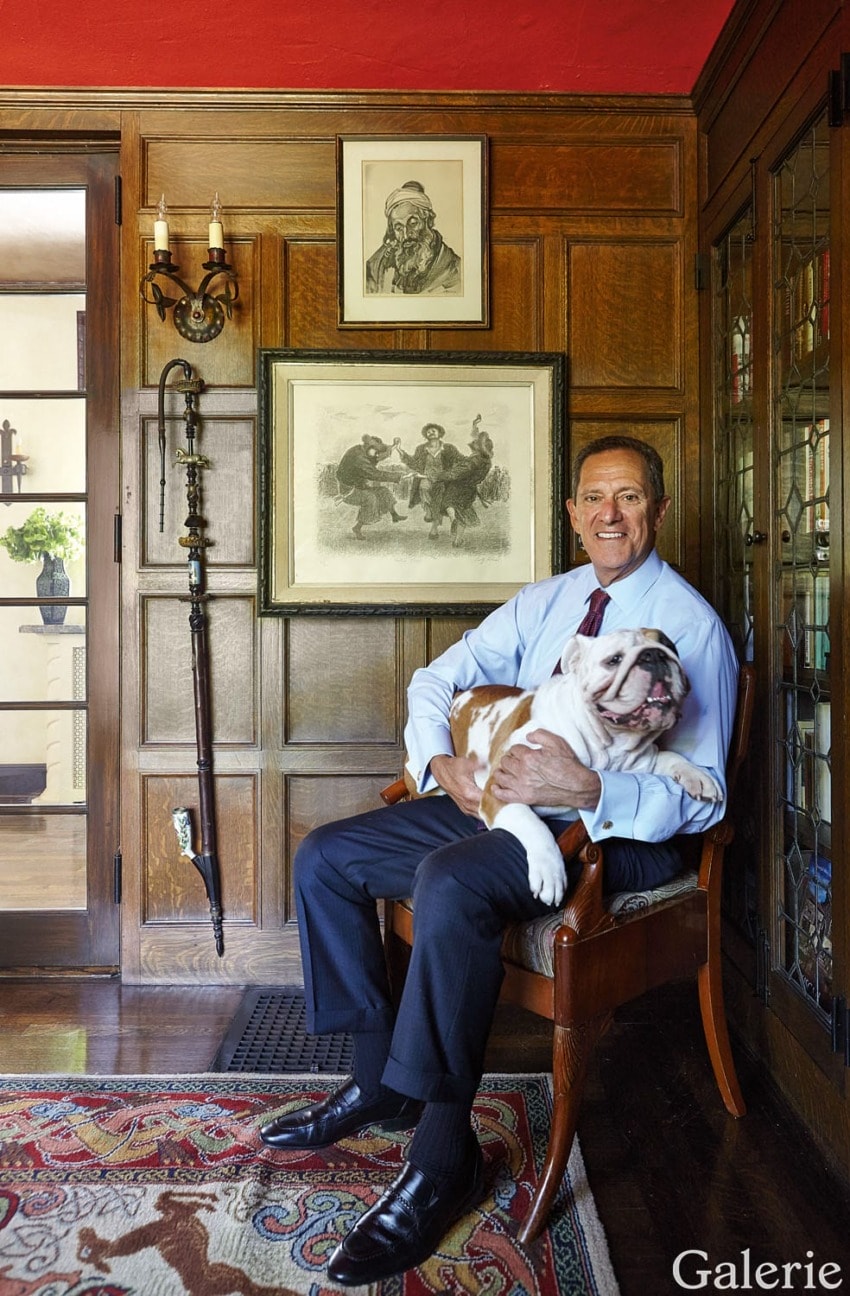 Man sitting in a wooden chair holding a bulldog, with artwork and a walking cane displayed on a wood-paneled wall in the background.