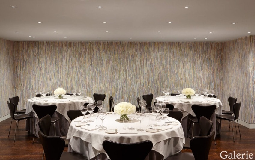 Elegant dining room with round tables, white tablecloths, set with wine glasses and floral centerpieces, modern art on walls.