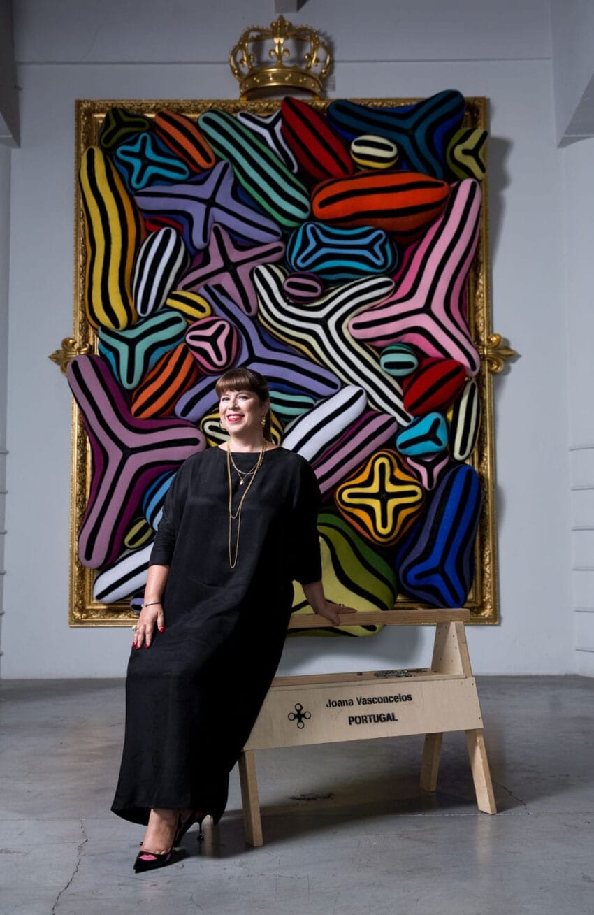 Person in a black dress sitting on a bench in front of a colorful abstract painting with bold patterns and shapes.