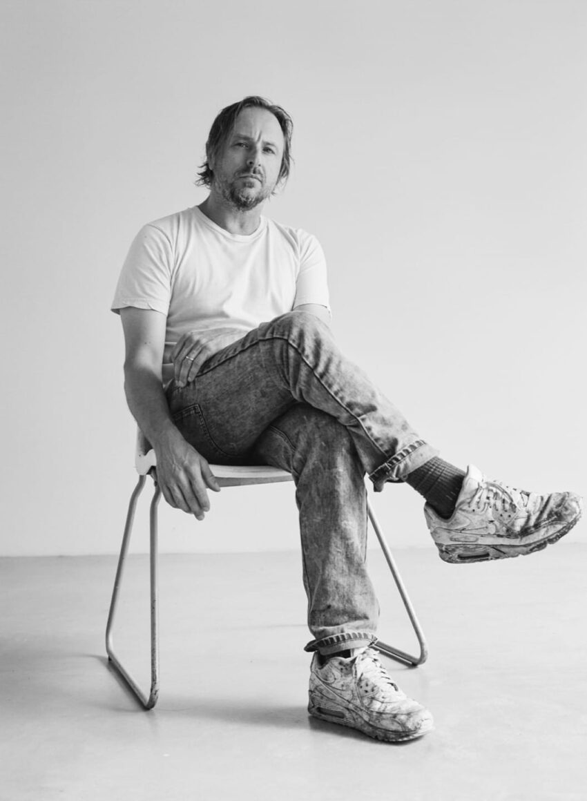 Man sitting on a chair in a simple room, wearing a white t-shirt and jeans, looking directly at the camera.
