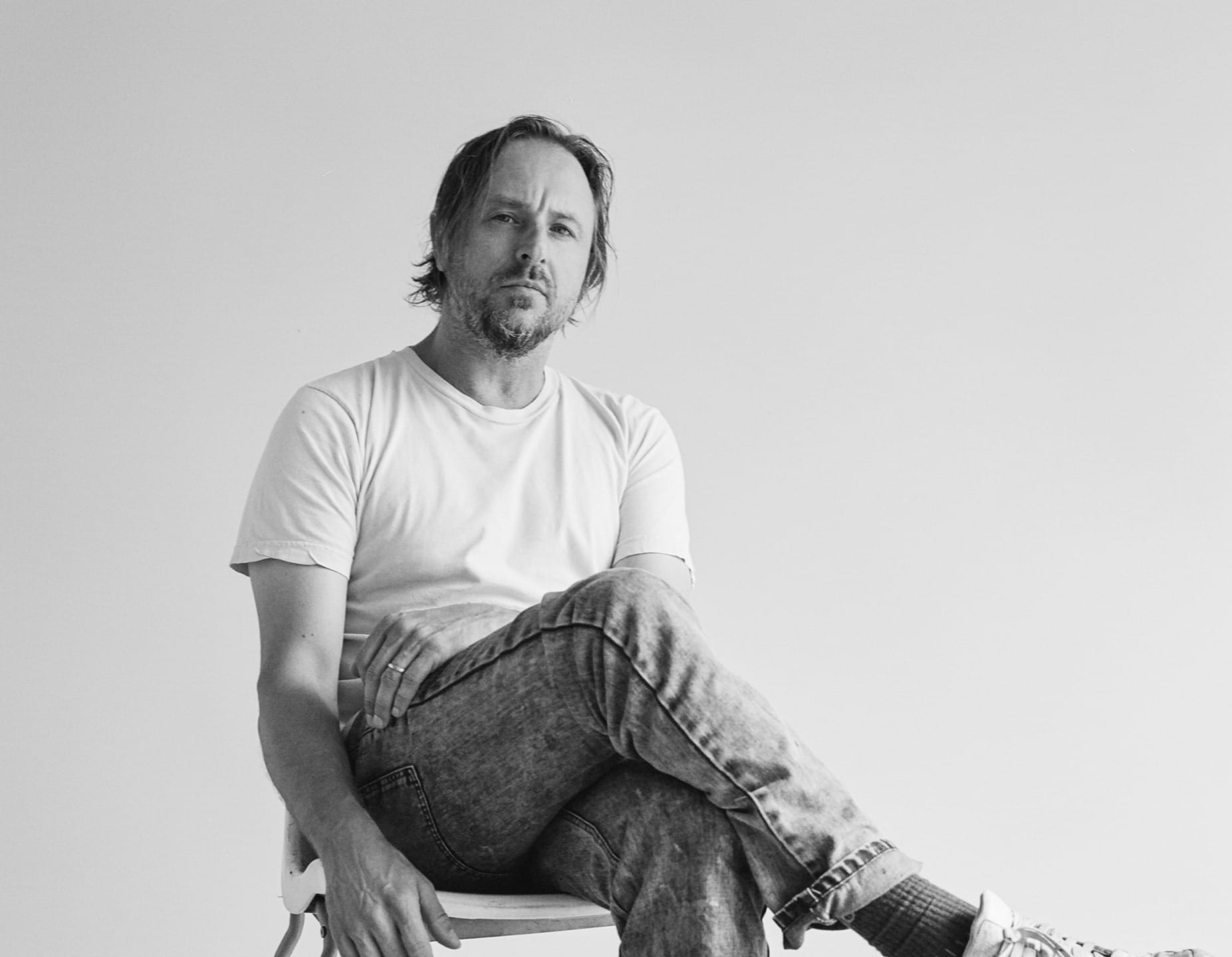 Man sitting on chair, wearing a white t-shirt and jeans, looking at the camera, in a black and white photo.