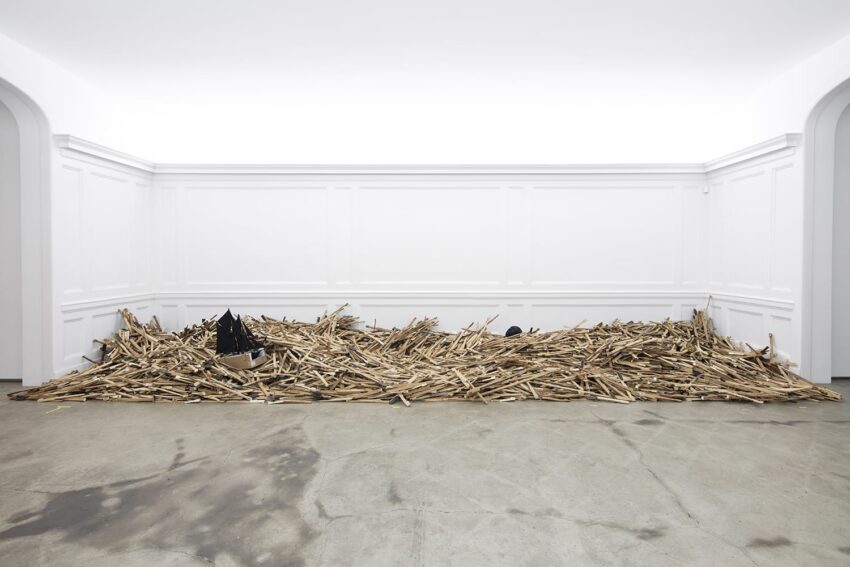 Large pile of wooden sticks with scattered debris in an empty white gallery space with high walls and concrete floor.