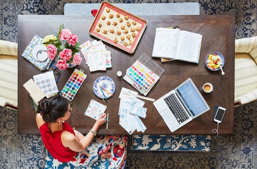 Woman painting at a table with art supplies, cookies, a laptop, and flowers on a patterned rug.