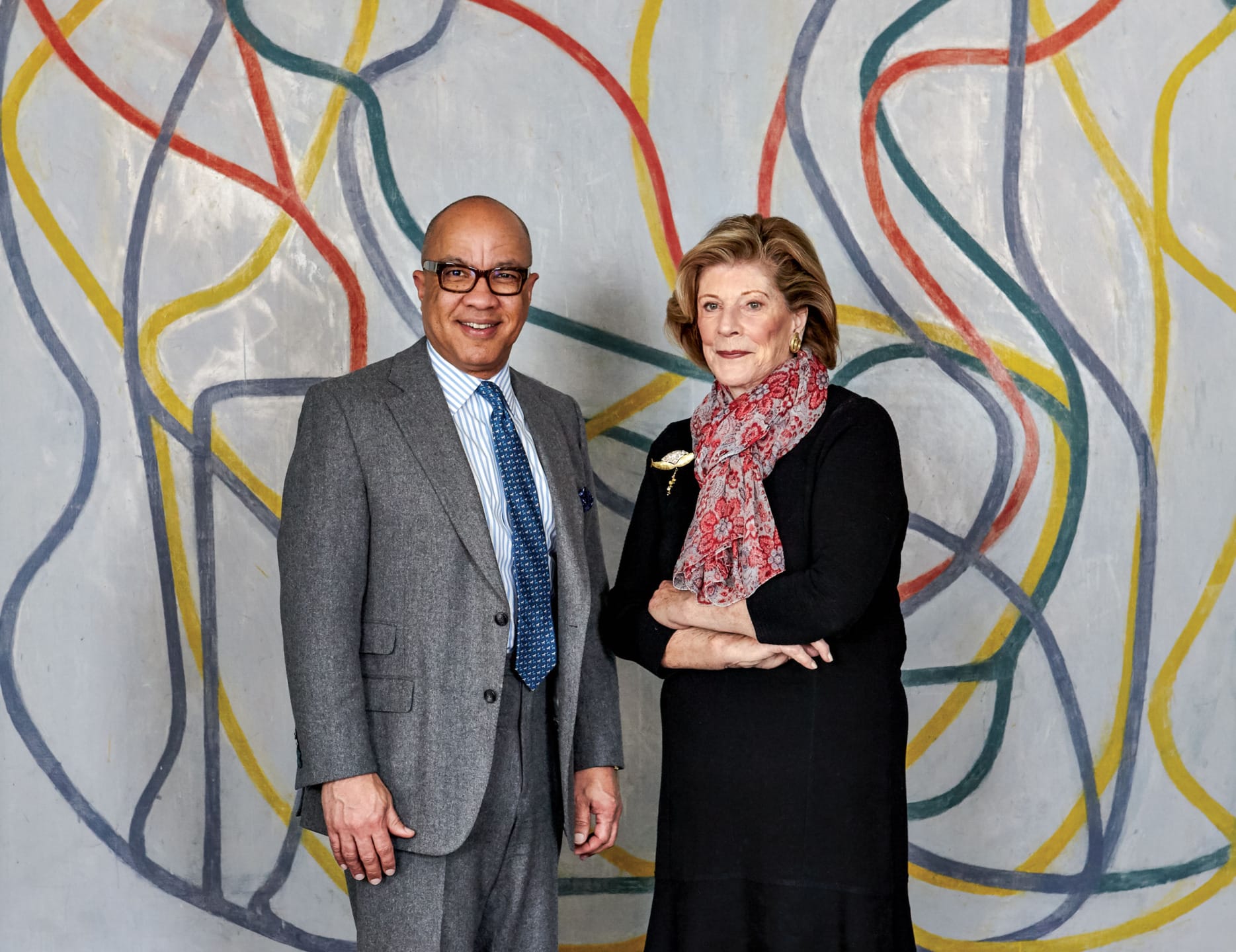Two people standing in front of a colorful abstract mural, one wearing a gray suit, the other in a black dress with a scarf.