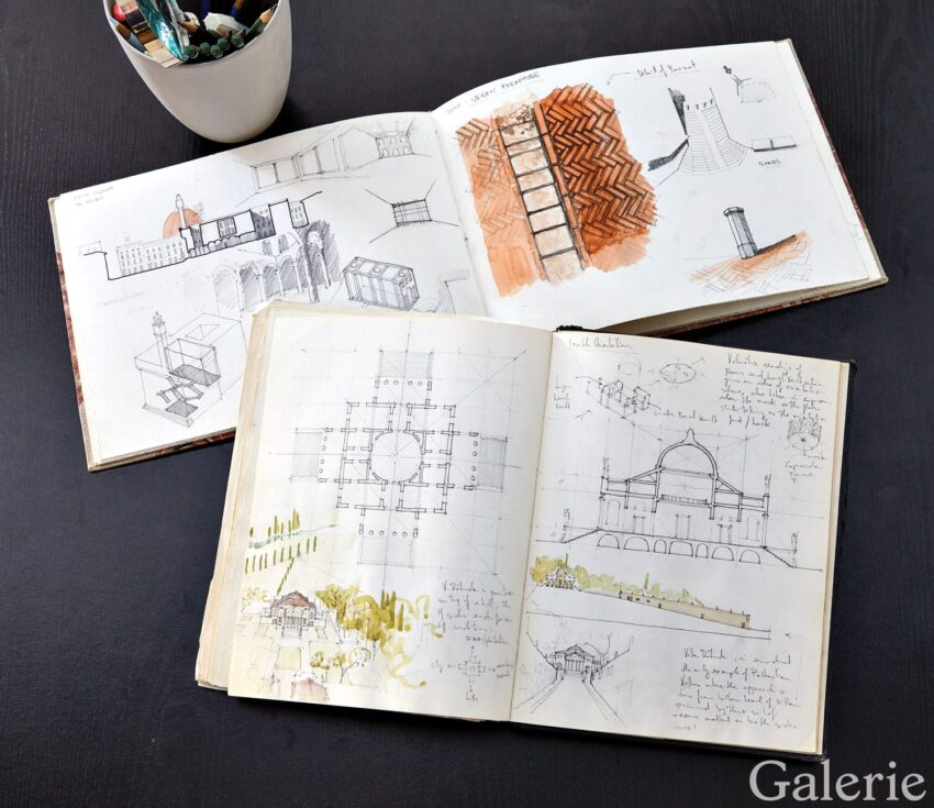 Open sketchbooks with architectural drawings and color illustrations on a dark table, next to a cup of colored pencils.