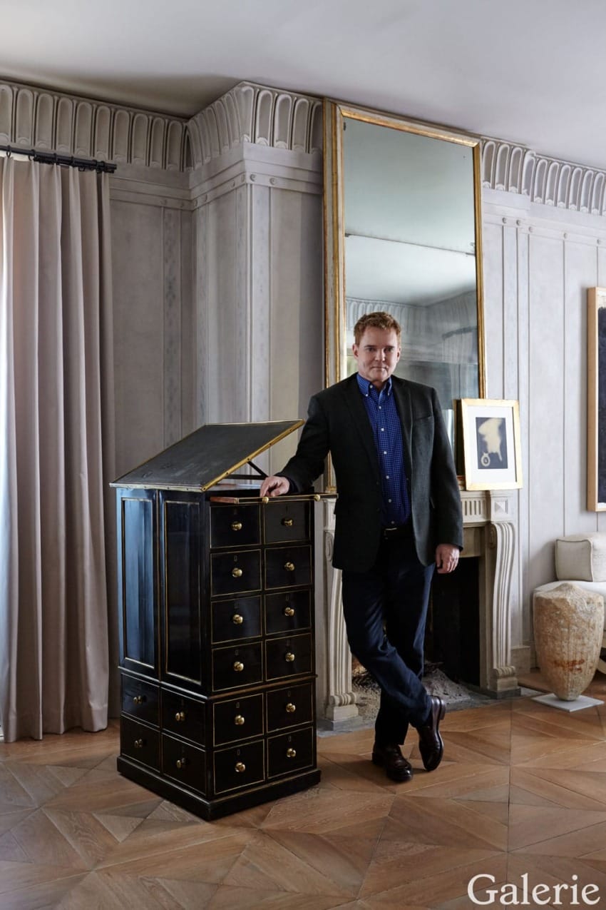 Person standing beside a black wooden cabinet in an elegant room with a tall mirror, art piece, and patterned curtains.