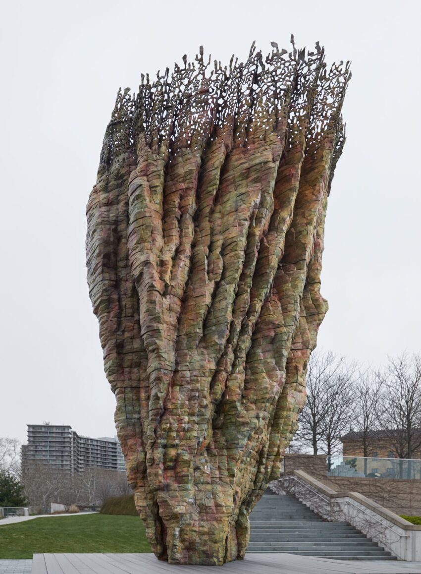 Large twisted sculpture resembling a colorful, textured seed pod with an open top, situated outdoors in a park setting.