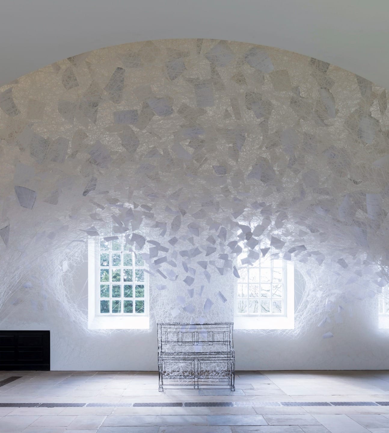 Artistic installation with white paper-like elements on a wall, surrounding windows, and a delicate metal structure on the floor.