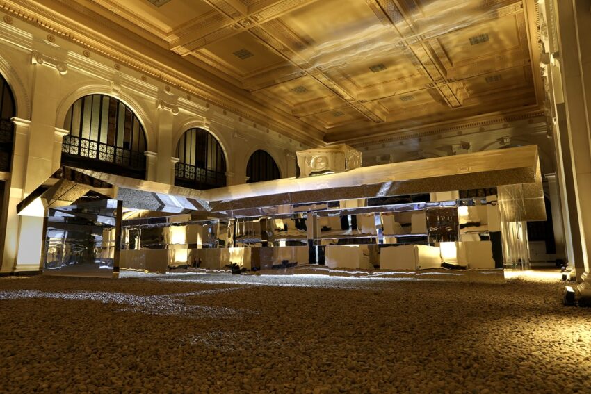 Large reflective installation in an ornate hall with arched windows and high ceilings, surrounded by small stones.