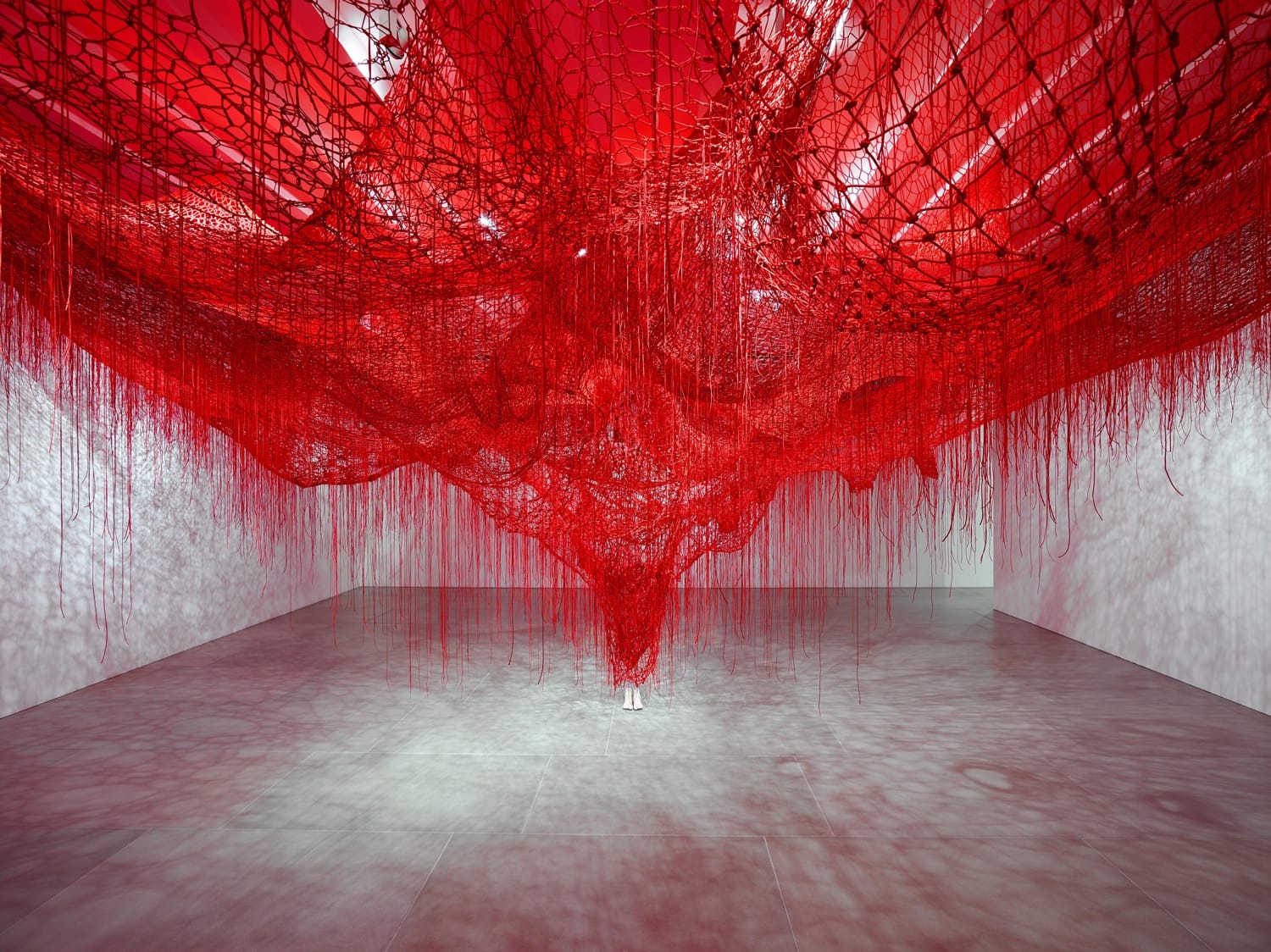 A large red yarn installation fills a modern gallery space with intricate patterns and shadows on the floor.