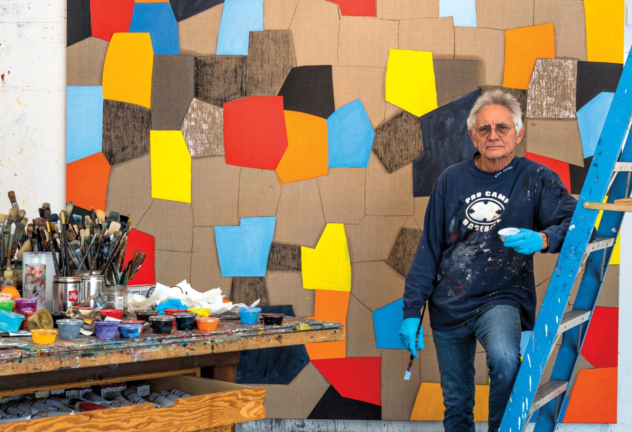 Artist in a studio with abstract geometric painting and art supplies on a table in the foreground.