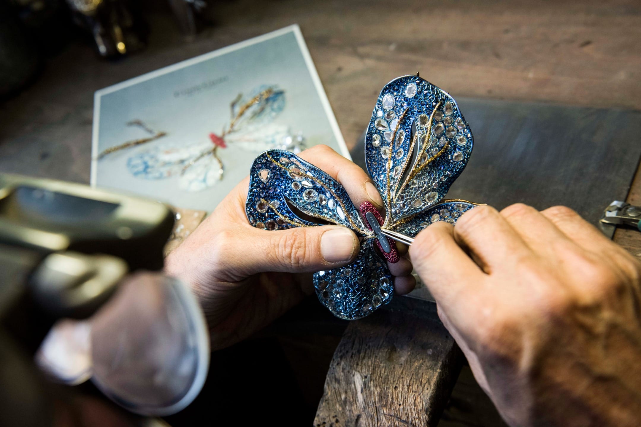 Craftsman intricately working on a detailed, bejeweled blue butterfly design at a wooden workbench.