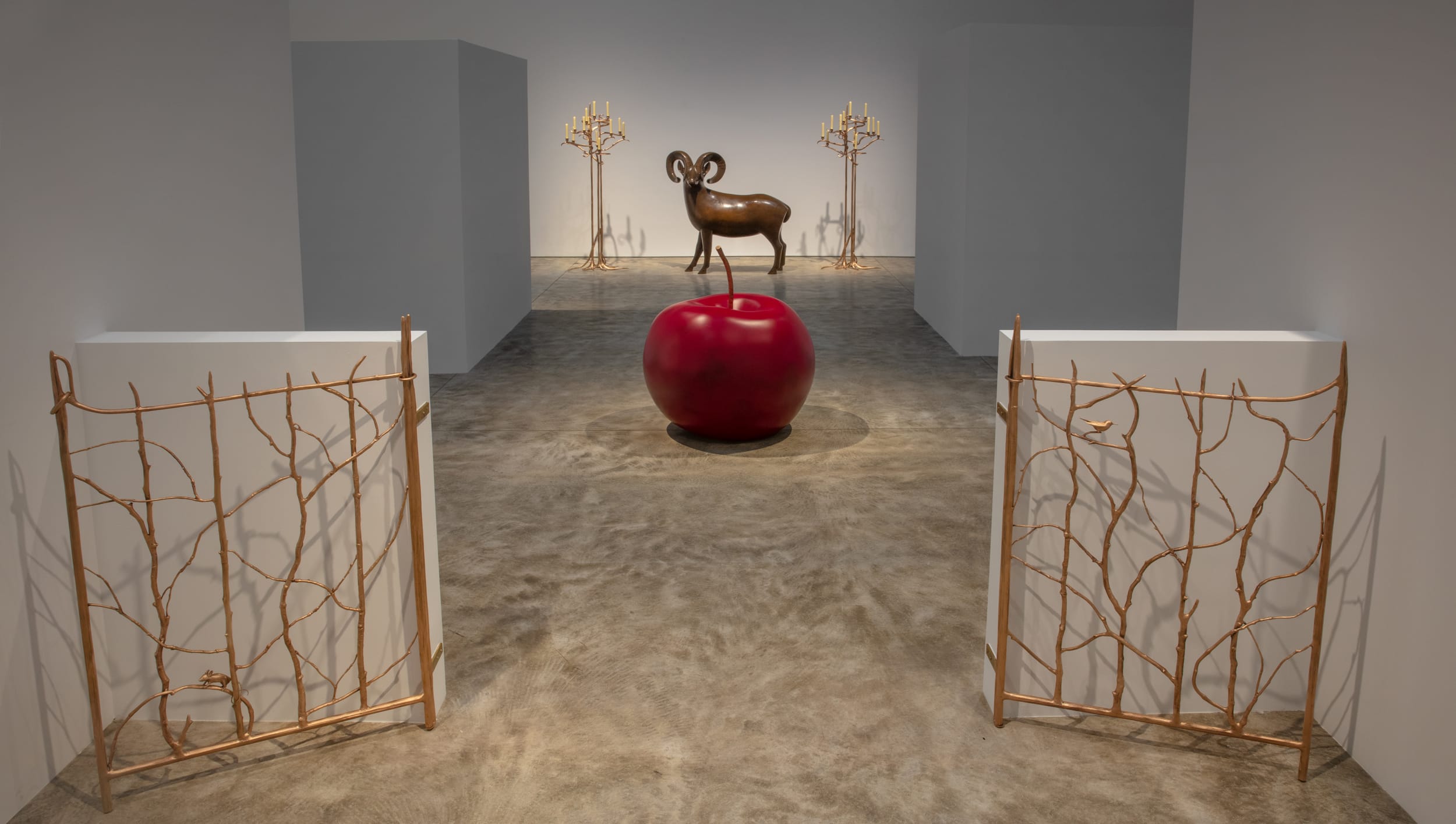 Art installation featuring a large red apple, metal gates, a ram sculpture, and artistic candelabras in a minimalist gallery setting.