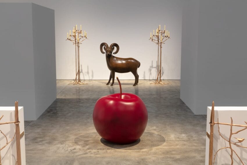 Art gallery with a large red apple sculpture in the foreground and a ram statue in the background, surrounded by candle holders.