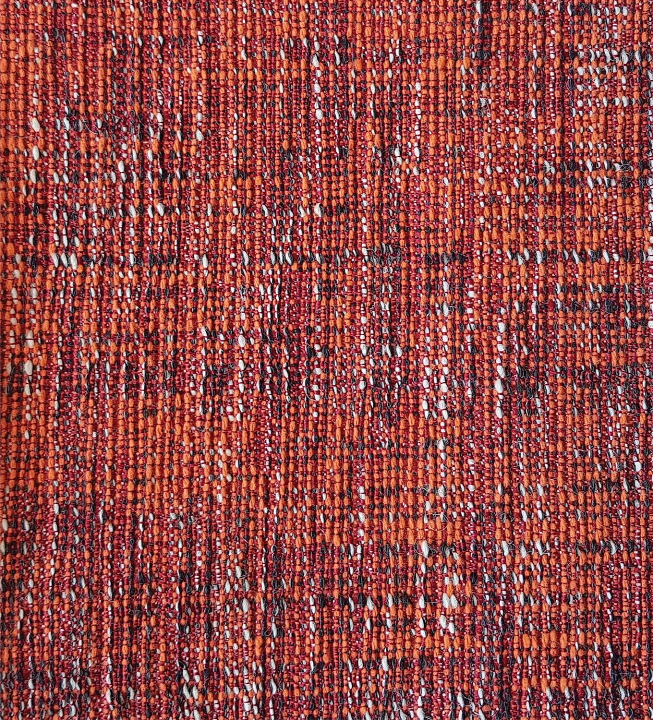 Close-up of a textured red and orange woven fabric with intricate patterns and various shades and flecks of white and black.