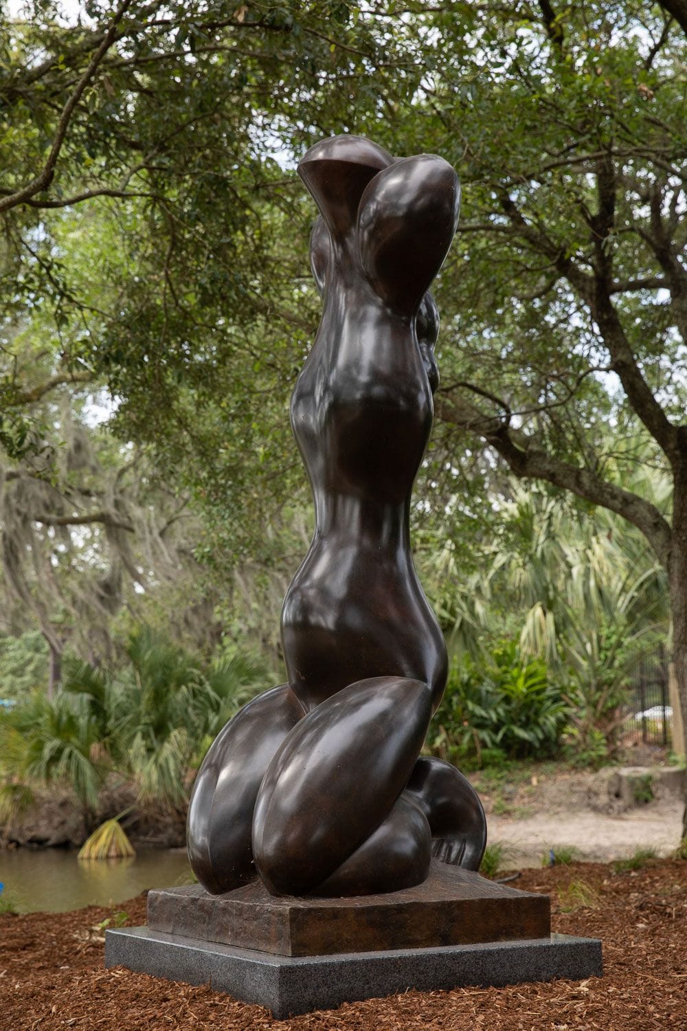 Bronze abstract sculpture of a humanoid figure in a garden setting with trees and greenery.